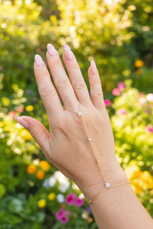 Chaîne de main dorée en acier inoxydable avec deux fleurs en zirconium et petits zirconiums le long de la chaîne, portée sur une main devant un fond floral coloré