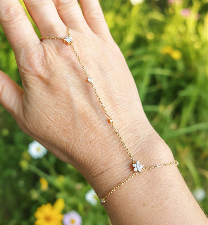 Chaîne de main dorée en acier inoxydable avec deux fleurs en zirconium et petits zirconiums le long de la chaîne, portée sur une main devant un fond floral coloré