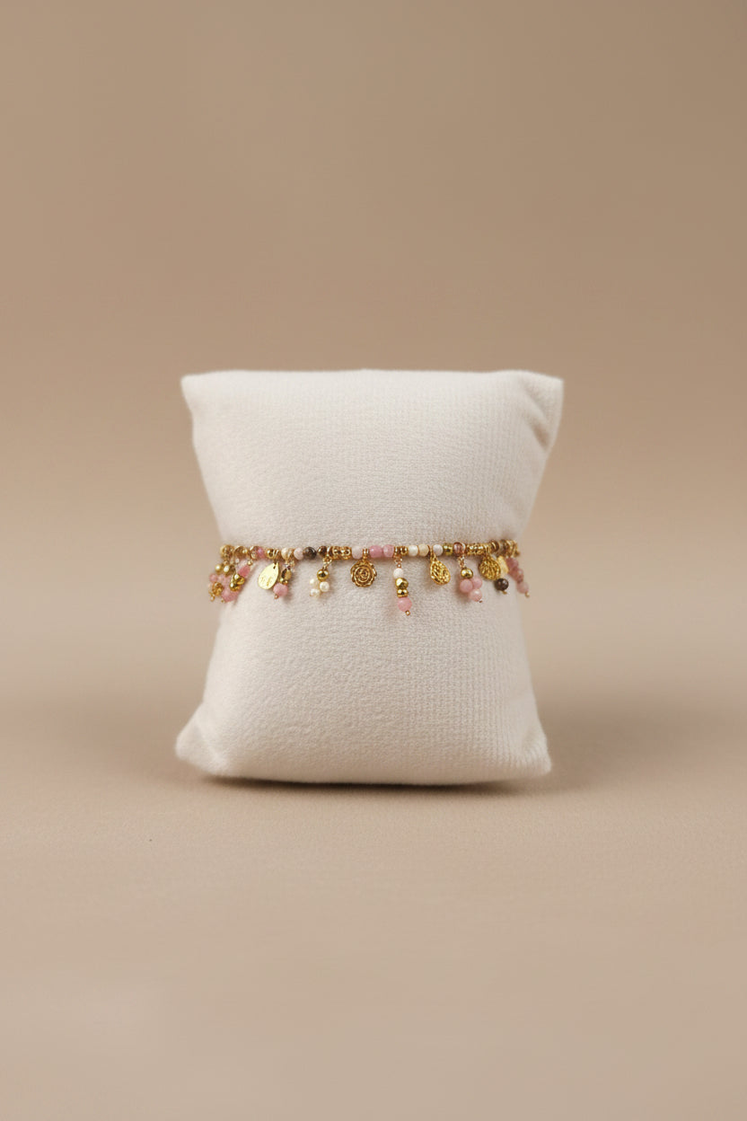 Bracelet quartz rose naturel et acier inoxydable doré avec breloques dorées, présenté sur coussin bijoutier blanc, fond beige – vue de face